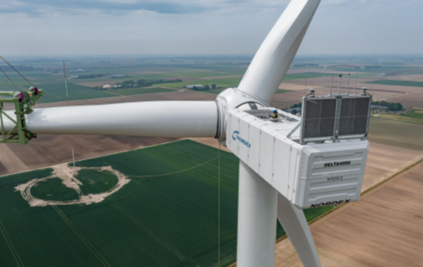Drone shot of the installation of a white Delta4000 wind turbine of the type N163/6.X. On the right site a crane lifting the last rotor blade. In the background green fields.