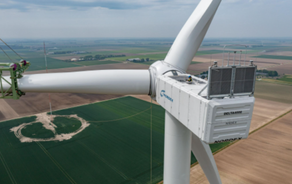 Drohnenaufnahme der Installation einer weißen Delta4000 Windturbine des Typs N163/6.X. Auf der linken Seite ein Kran, der das letzte Rotorblatt anhebt. Im Hintergrund grüne Felder.